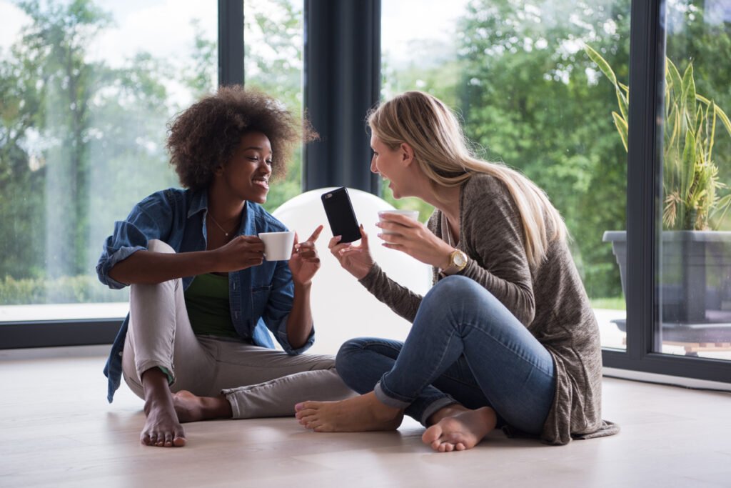 Two young laughing multiethnic women sit on the floor near the window and enjoy while drinking coffee using smartphone in luxury home